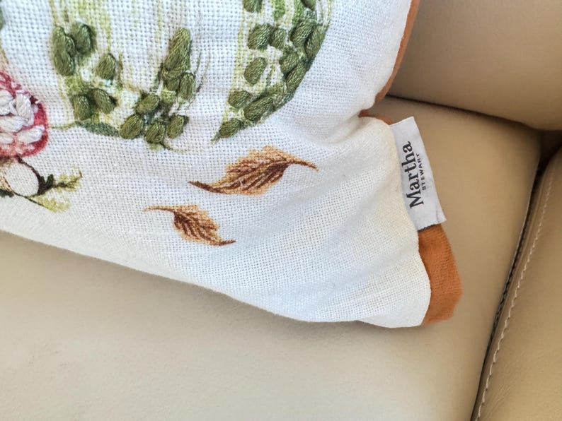 Pu&ograve; includere: Primo piano di un cuscino decorativo con un motivo floreale e di foglie. Il cuscino presenta uno sfondo bianco con accenti ricamati verdi, rosa e marroni. Il cuscino ha un'etichetta Martha Stewart.