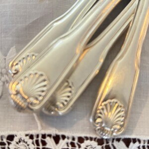 May include: Close-up of several silver knives with a shell design on the handles. The knives are arranged diagonally on a white tablecloth with a decorative lace border. The blades are shiny and reflect light.