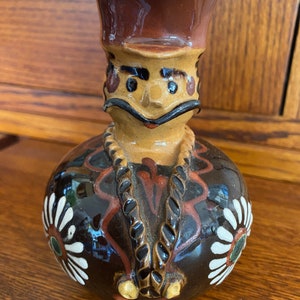 May include: A brown ceramic pitcher with a whimsical face, a brown hat, and white daisy designs. The pitcher has a brown rope-like design around the neck.