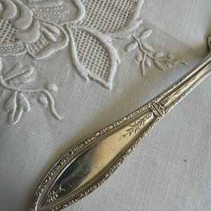 May include: A silver, ornate, decorative, flatware piece with a floral design. The piece is lying on a white linen tablecloth with embroidered flowers.