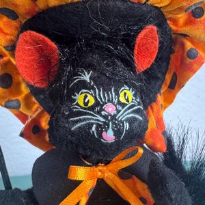 May include: A black cat figurine wearing an orange and black polka dot bonnet and a black dress with an orange ribbon bow. The cat has yellow eyes, red inner ears, and a pink nose. It holds a small broom made of light-colored straw fibers.