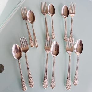 May include: A set of eight silver-toned metal utensils consisting of four forks and four spoons. Each piece features a polished finish and a decorative floral pattern embossed on the handle base. The items are arranged in pairs on a smooth surface.