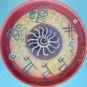 May include: A decorative ceramic plate with a maroon rim and a central design featuring a sunburst and stylized figures in teal and white. The plate has a textured, speckled background and the artist's signature.