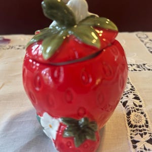 May include: A ceramic strawberry-shaped jar with a lid, featuring a glossy red finish and green leaves. A smaller strawberry with a white flower is attached to the base. The jar is likely for kitchen or home decor.