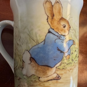 May include: A white ceramic mug featuring a detailed illustration of Peter Rabbit in a blue jacket. The artwork depicts the rabbit in a field with green foliage and a yellow background. The mug has a curved handle.