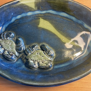 May include: A blue and green ceramic oval dish with two raised crab designs. The dish is glazed and has a glossy finish.