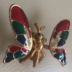 Puede incluir: Un broche de mariposa de tono dorado con alas de esmalte rojo, verde y azul. Las alas est&aacute;n decoradas con diamantes de imitaci&oacute;n transparentes. La mariposa tiene un cuerpo y antenas detalladas.