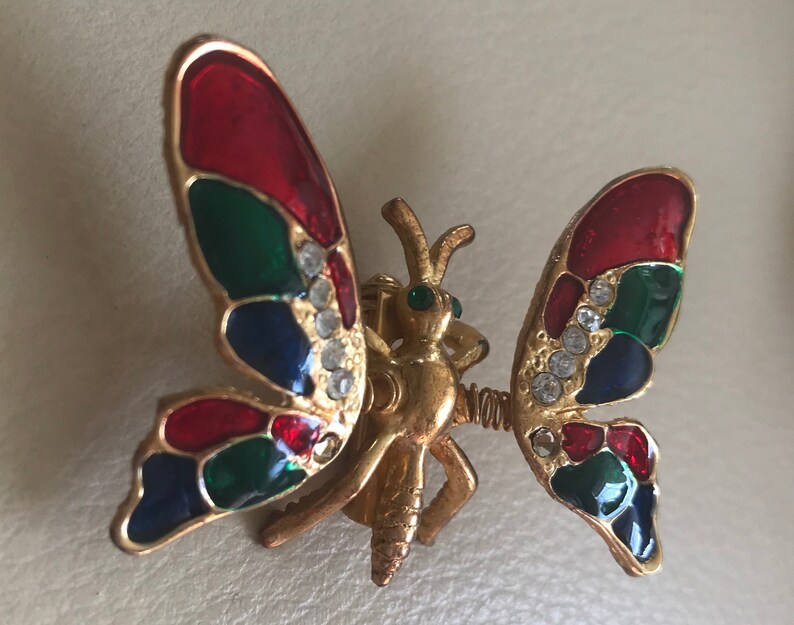 Puede incluir: Un broche de mariposa de tono dorado con alas de esmalte rojo, verde y azul. Las alas est&aacute;n decoradas con diamantes de imitaci&oacute;n transparentes. La mariposa tiene un cuerpo y antenas detalladas.