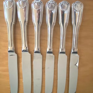 May include: A set of six silver-toned dinner knives arranged in a row. Each knife features a polished metallic finish and a decorative shell-shaped motif at the top of the handle. The handles have a smooth, tapered design leading to a sharp blade.