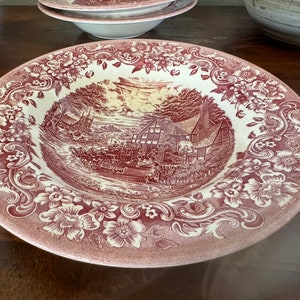 May include: A large, red and white ceramic bowl with a floral design around the rim. The bowl features a vintage scene of a village with a church and houses.