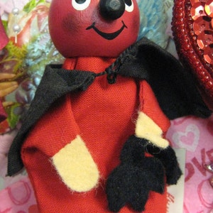 May include: A red felt puppet with a black hat and cape. The puppet has a black nose and a wide, toothy grin. The puppet is wearing a red felt dress and has white felt hands.