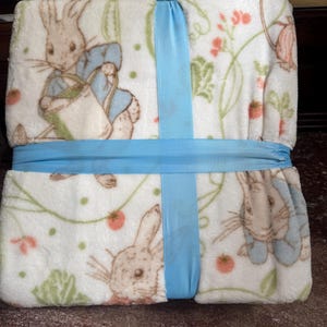 May include: A soft, white baby blanket featuring illustrations of Peter Rabbit with a light blue ribbon wrapped around it. The blanket has a repeating pattern of Peter Rabbit, flowers, and greenery.