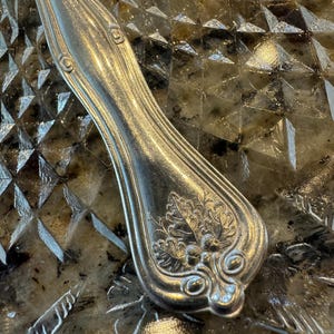 May include: Close-up of the ornate, silver handle of a vintage fork. The handle is decorated with a floral design and has a polished finish.