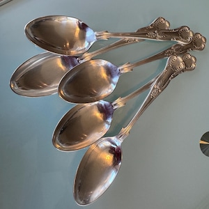May include: Five silver spoons with ornate, floral-like detailing on the handles. The spoons are arranged diagonally, reflecting light. The spoons have a classic, vintage aesthetic, suggesting they are antique or vintage silverware.