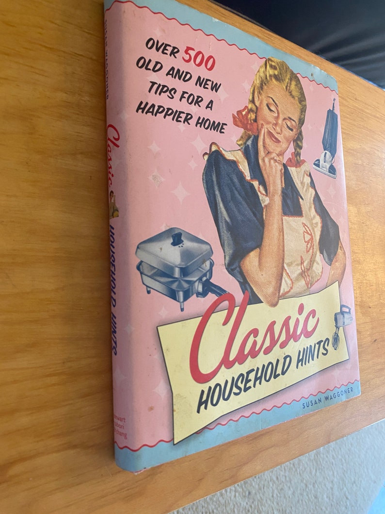 Pode incluir: Uma capa de livro rosa e azul com o t&iacute;tulo "Classic Household Hints" e o texto "Over 500 Old and New Tips for a Happier Home". A capa apresenta uma mulher com um vestido azul e branco, uma m&aacute;quina de waffle e um misturador.