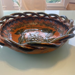 May include: A brown ceramic serving dish with a woven rim and a green and brown design. The dish is decorated with a floral pattern.