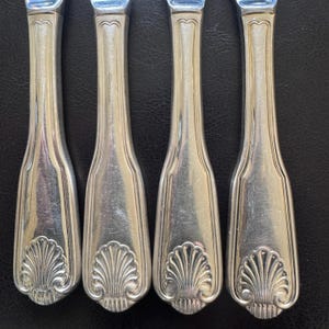 May include: Four silver-toned metal cutlery handles arranged vertically. Each handle features a smooth, tapered shaft with a raised, fan-shaped shell motif at the base and a subtle, curved decorative line detail at the top near the neck.