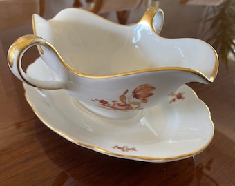 May include: A white porcelain gravy boat with a gold rim and floral design. The boat is on a matching saucer.