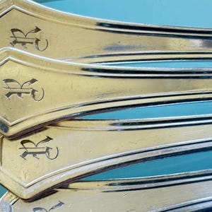 May include: Close-up of several gold-colored silverware pieces, possibly forks or spoons, with a decorative design. Each piece has a stylized letter "R" engraved on the handle. The background is a light teal color.