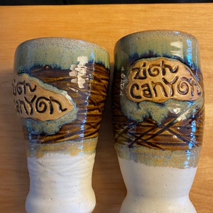 May include: Two brown and green ceramic mugs with white bases. Each mug has the words "Zion Canyon" written on it in brown.