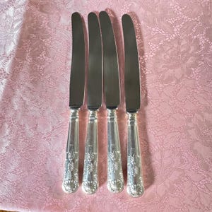 May include: Four silver knives with ornate handles are arranged on a pink floral tablecloth. The knives have a classic design with a shiny, reflective blade and detailed handle. The handles feature a decorative pattern.
