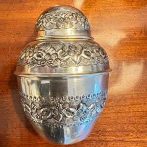 May include: A silver-colored metal container with a rounded lid, featuring embossed floral patterns and textured detailing around the top and middle sections. The object has a smooth, polished finish and a bulbous, egg-like shape with a secure lid.
