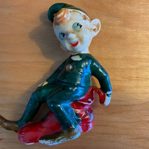 May include: A ceramic figurine of an elf wearing a green suit and a green hat, sitting on a red and white sled. The elf has a white face with painted features and a mischievous expression.