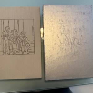 May include: A vintage hardcover book with a beige cover and a black and white illustration of three people on the front. The book is closed and the spine is visible. The book is next to a silver-colored book with a textured cover.