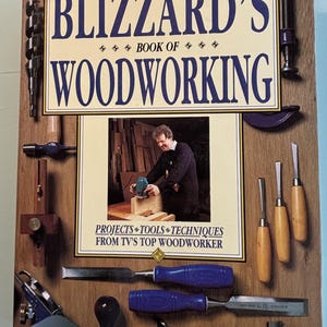 May include: A book titled "Blizzard's Book of Woodworking" with a photo of a man using a power tool. Various woodworking tools, including chisels, drills, and a plane, are arranged around the book on a wooden background.