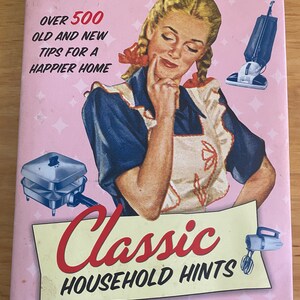 Pode incluir: Uma capa de livro de estilo vintage com fundo rosa e uma mulher em um vestido azul e branco. O t&iacute;tulo do livro &eacute; "Classic Household Hints" e o subt&iacute;tulo &eacute; "Over 500 Old and New Tips for a Happier Home".