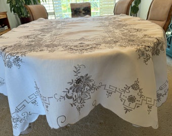 May include: White linen tablecloth with a scalloped edge and intricate embroidered floral designs in gray.