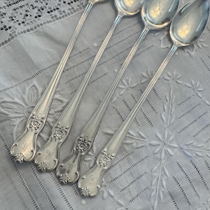 May include: Four silver spoons with ornate floral and scroll detailing on the handles, each with a monogram. The spoons are arranged diagonally on a light blue embroidered cloth.