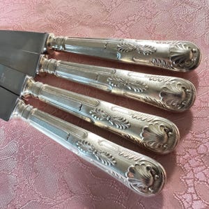 May include: Close-up of four silver knives with ornate handles. The handles feature a detailed, symmetrical design with floral and scrollwork patterns. The blades are smooth and shiny, reflecting the light. The knives are arranged diagonally on a pink patterned surface.