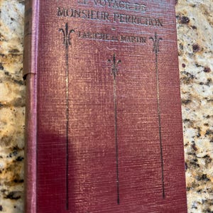 May include: A burgundy-colored book with the title "Le Voyage de Monsieur Perrichon" and the author "Labiche & Martin" printed on the cover.
