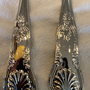 May include: Close-up of the ornate handles of two silver spoons. The handles are decorated with a floral design and a shell motif.