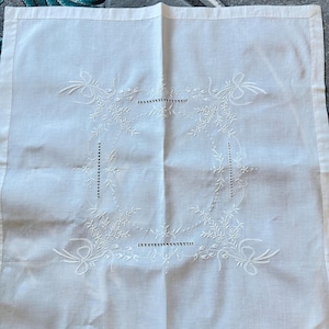 May include: White linen tablecloth with intricate embroidered floral and ribbon design. The embroidery forms a square frame with delicate leaves and dotted lines. The edges are neatly hemmed, creating a classic and elegant look.