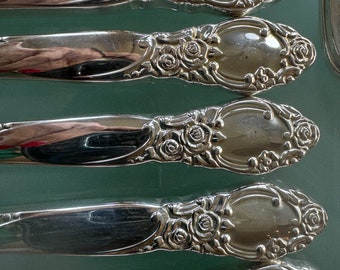 May include: Five silver-plated dinner forks with ornate rose designs. The forks are arranged in a stack, with the handles facing up.