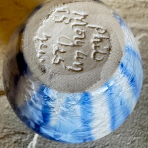 May include: A handmade ceramic bowl with a blue and white crystalline glaze. The bowl's exterior features a striking pattern of blue and white streaks, while the base has stamped text. The bowl is a unique piece of art.