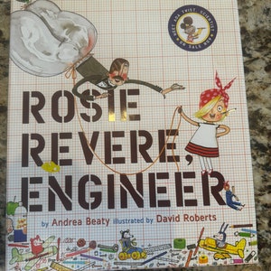 May include: A hardcover children's book titled "Rosie Revere, Engineer" by Andrea Beaty, illustrated by David Roberts. The cover features illustrations of characters and engineering tools on a graph paper background. The book is a New York Times bestseller.