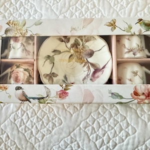 May include: A boxed tea set featuring floral and bird designs. The set includes four teacups, saucers, and a serving plate. The box is white with pink and green floral and bird illustrations.