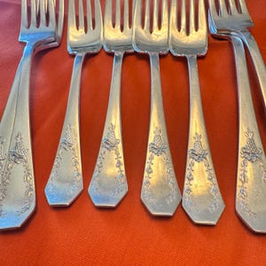 May include: A set of six silver-toned metal forks arranged in a row. Each fork features a polished finish and a decorative embossed pattern on the handle, consisting of a small basket motif at the top with trailing floral vines extending downward.