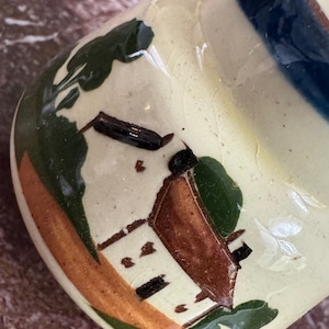 May include: A ceramic vase with a cream-colored base and a painted landscape design. The design features a brown building, green foliage, and accents of orange and blue. The vase has a rounded body and a slightly flared rim.