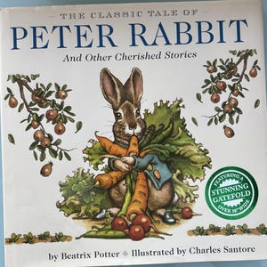 May include: A book cover featuring a brown rabbit holding carrots and other vegetables. The title is "The Classic Tale of Peter Rabbit and Other Cherished Stories" with an illustration of a rabbit holding carrots. The book features a stunning gatefold over 38 inches wide.