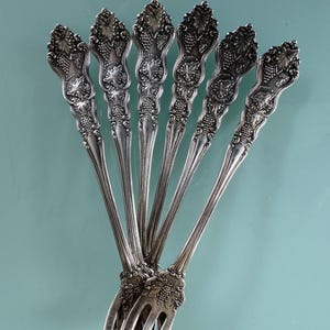 May include: Set of six silver-plated cocktail forks with ornate grapevine designs. The forks are arranged in a fan shape, with the tines pointing downwards.