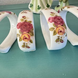 May include: White ceramic napkin rings with a floral rose design in shades of red and yellow. The rings are curved and have a glossy finish. The background is a light teal color.