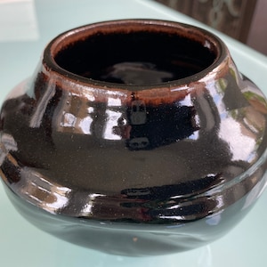 May include: A black ceramic vase with a glossy finish and a slightly rounded shape. The vase has a dark brown rim.
