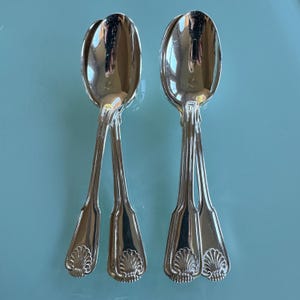 May include: Two silver serving spoons with a shell design on the handle. The spoons have a reflective surface and are arranged against a light blue background. The spoons are likely vintage or antique.