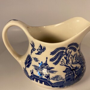 May include: A white ceramic creamer with a blue and white willow pattern. The creamer has a handle and a spout.