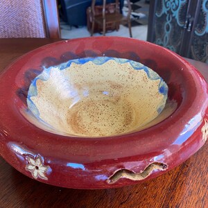 May include: A large, red ceramic bowl with a yellow interior and blue and brown accents. The bowl has a wavy rim and decorative floral and wavy designs.