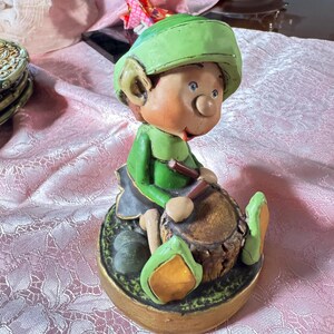 May include: A whimsical figurine of a seated leprechaun playing a drum. The leprechaun wears a green hat, shirt, and shoes, with a gray skirt. The drum is brown, and the base is gold.